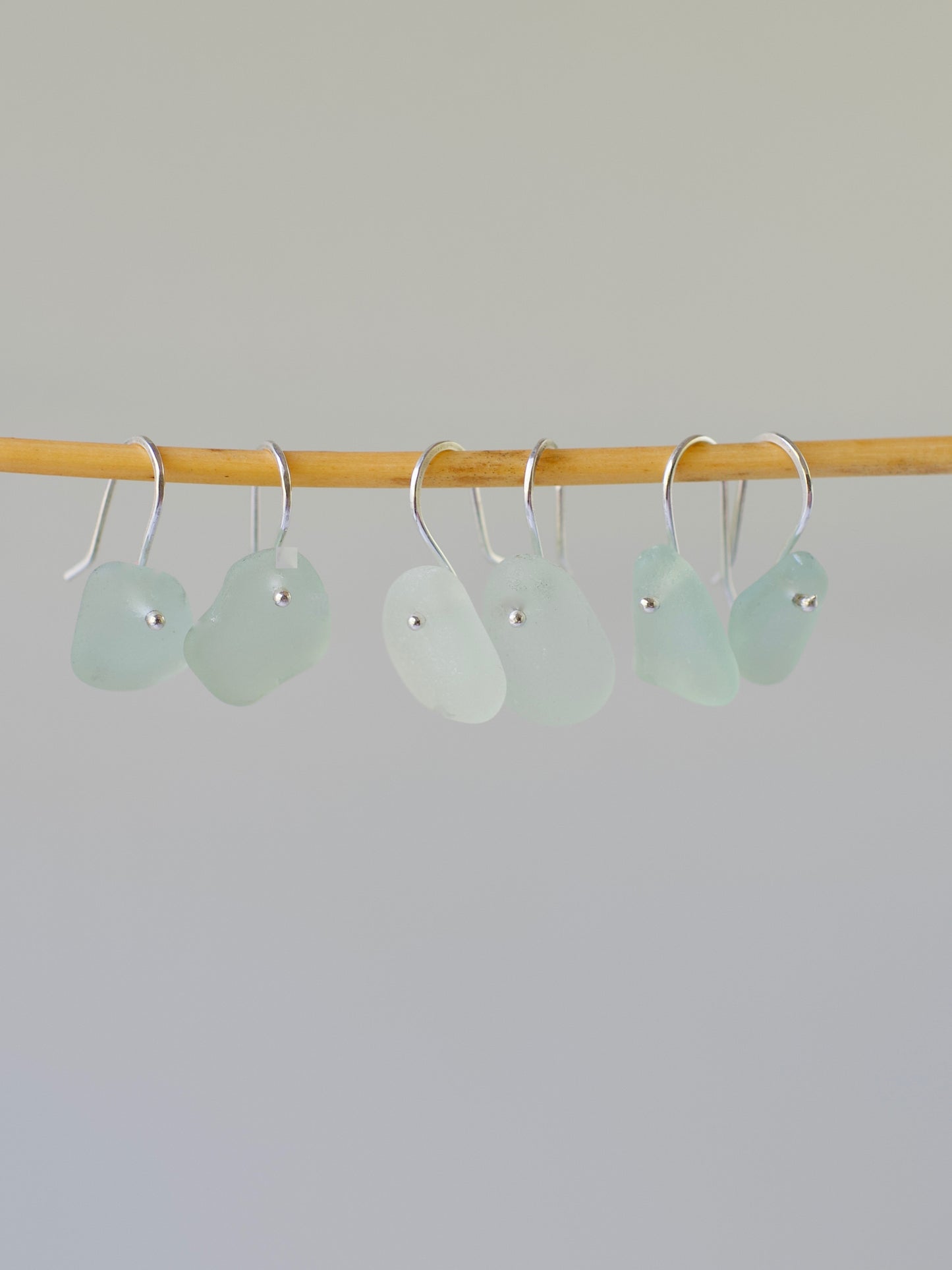 three pairs light aqua beach glass french ear wire earrings on a wooden rod against a gray background