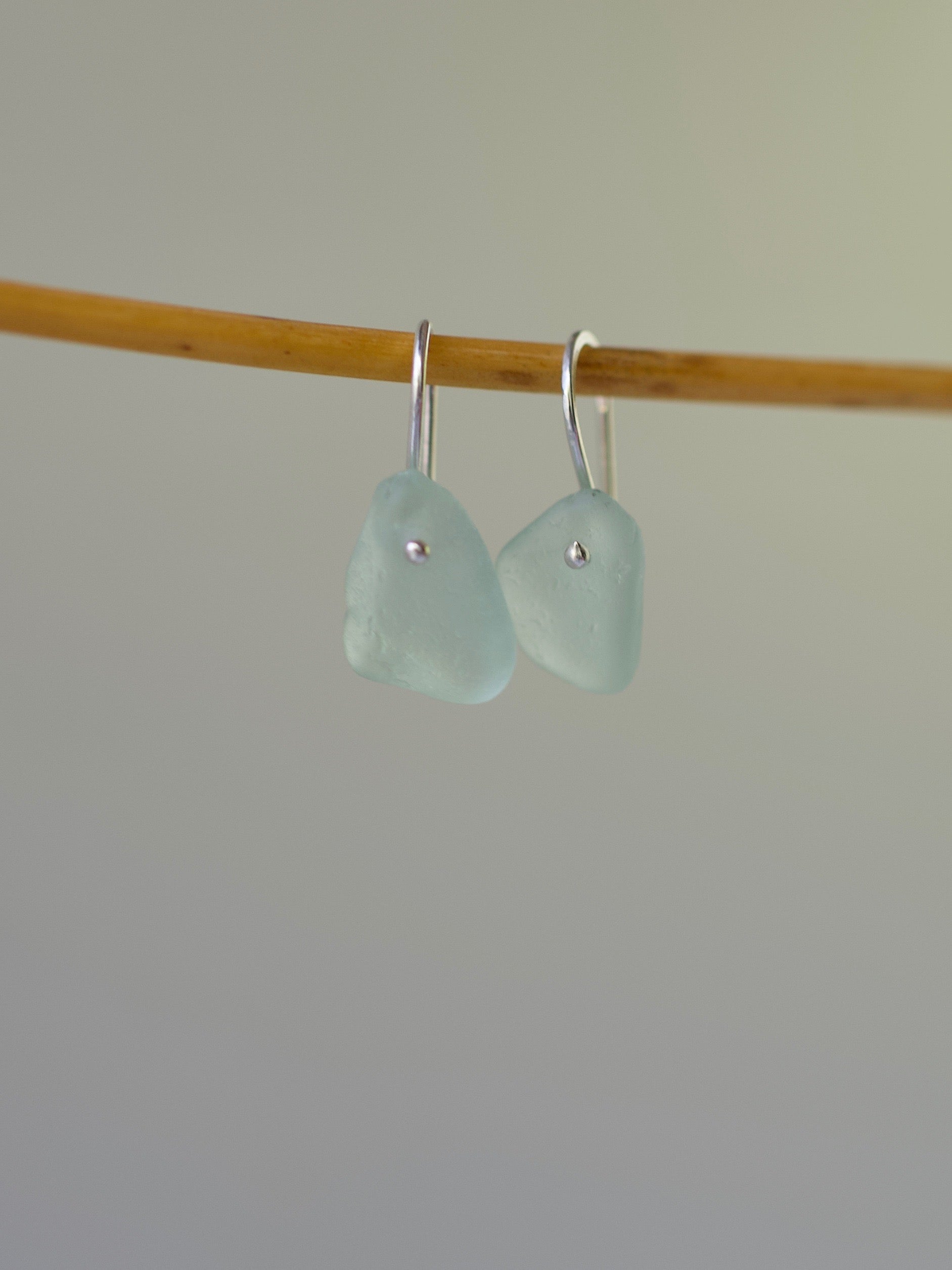 Pair of light blue sea glass earrings on a wooden stick against a plain background