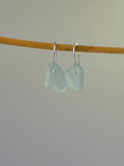 Pair of light blue sea glass earrings on a wooden stick against a plain background
