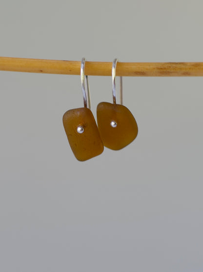 Pair of amber-colored sea glass earrings on a bamboo stick against a gray background