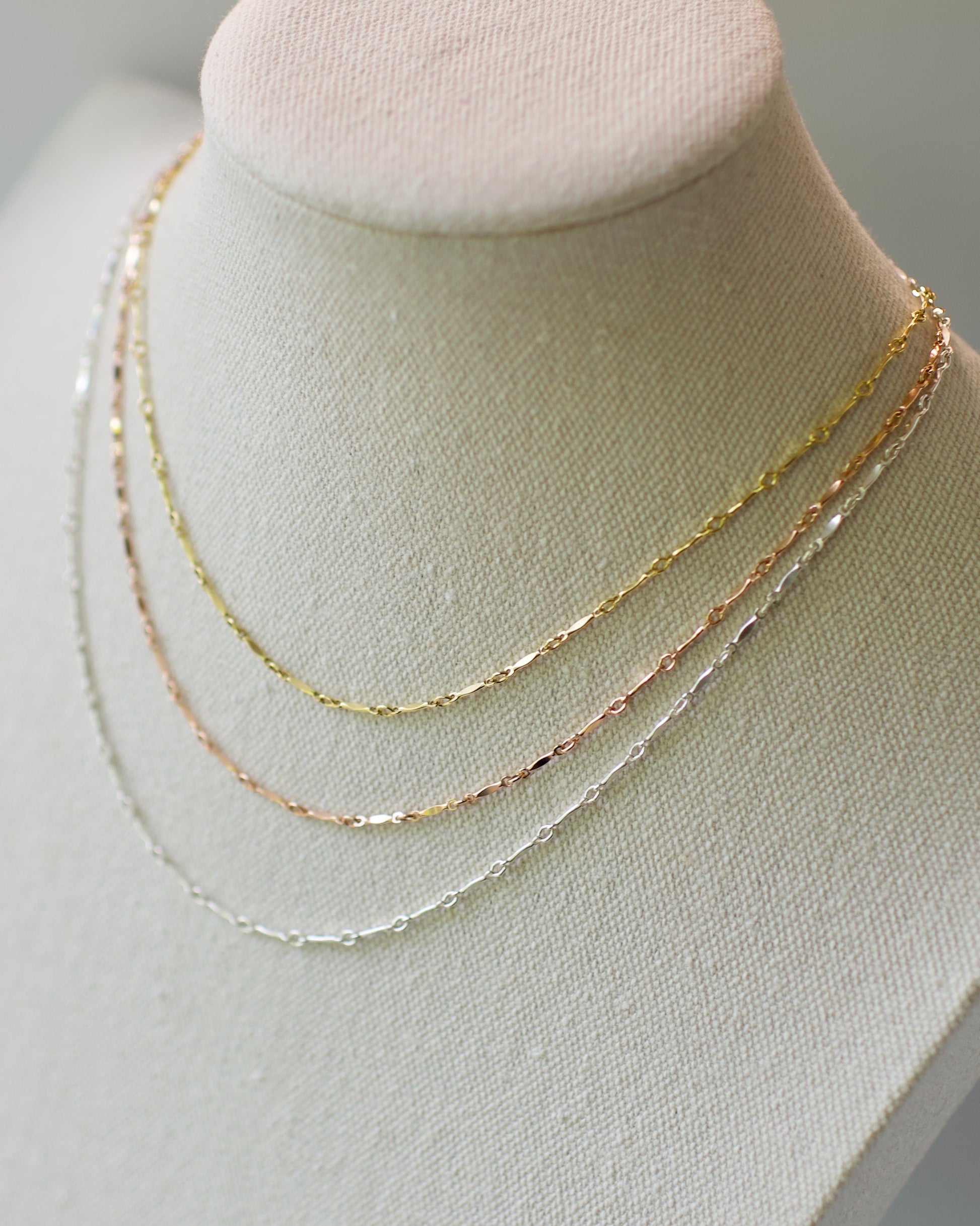 Three necklaces in a dapped bar and link chain style, yellow 14k usa gold fill, rose 14k usa gold fill, ,92 on a mannequin bust against a light background sterling silver